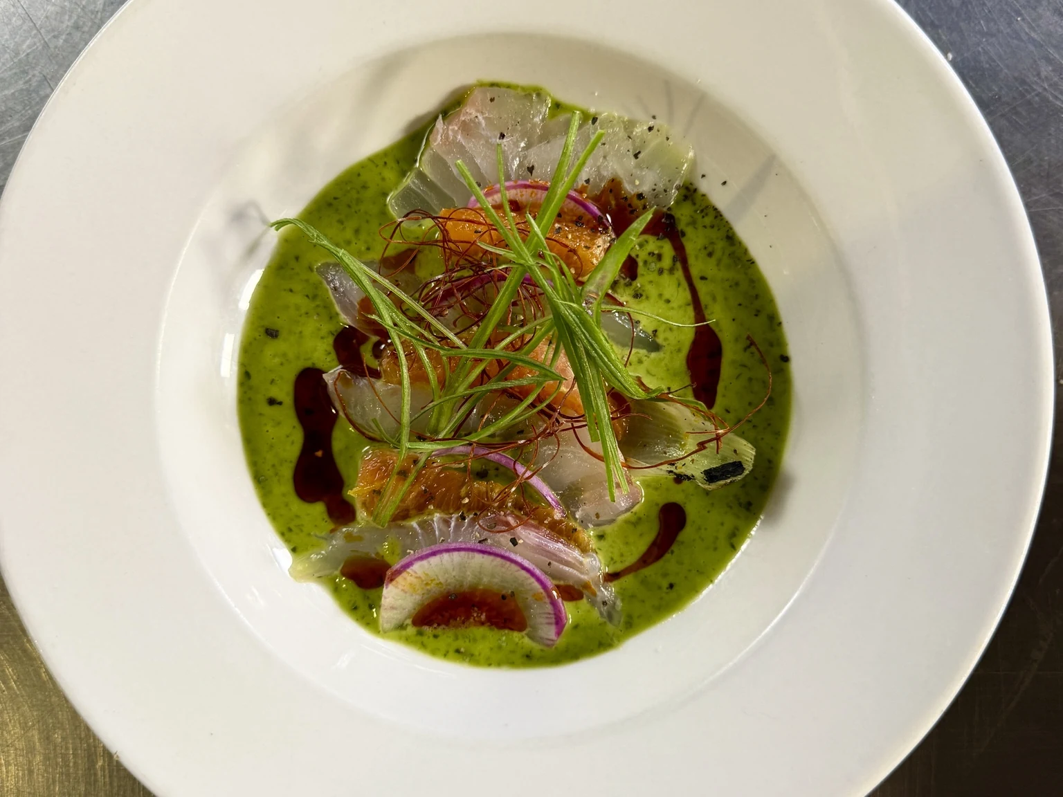 A plated seafood dish with green sauce, red onion, and herbs